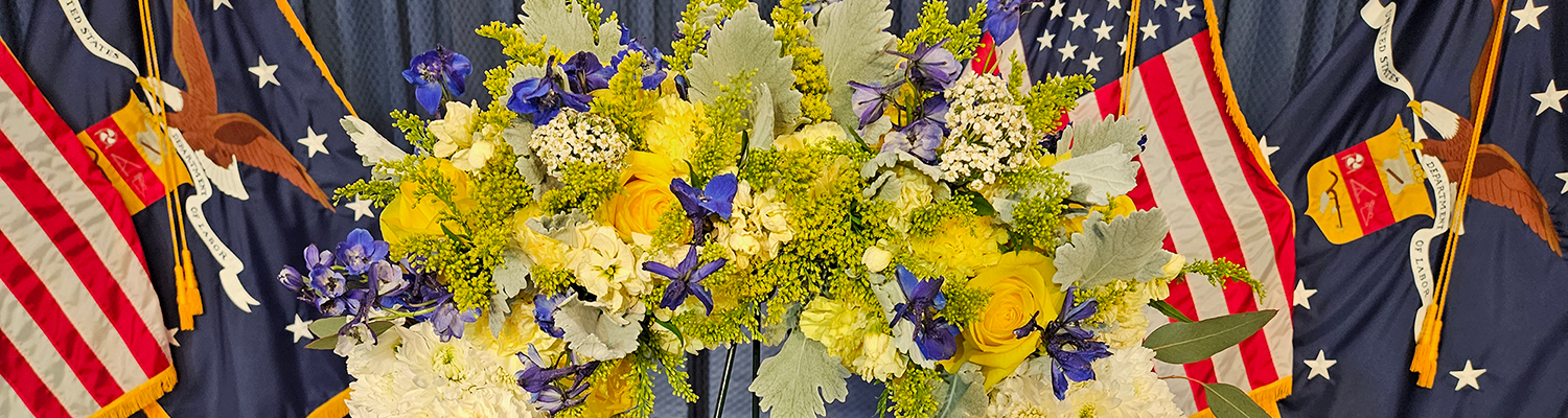 A wreath with white, yellow, and purple flowers standing in front of United States and Department of Labor flags